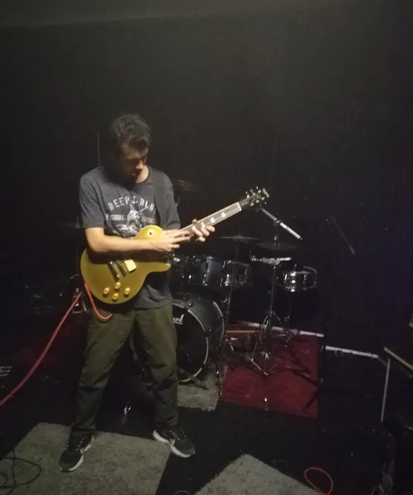Sinan playing a gold electric guitar in a rehearsal studio, drum kit behind him